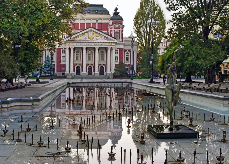 Stunning view of Ivan Vazov Theatre with reflection in a fountain pool in Sofia, Bulgaria, surrounded by trees.